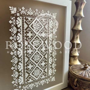 K&ouml;nnte beinhalten: Ein wei&szlig;er gerahmter Druck mit einem geometrischen Muster in Wei&szlig; auf beigefarbenem Hintergrund. Das Muster ist ein traditionelles Volksdesign mit einem sich wiederholenden Motiv aus Quadraten und Rauten.