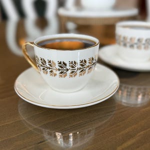 May include: A white teacup and saucer set with gold trim and a geometric gold pattern. The cup is filled with dark liquid, likely coffee or tea. The set is on a reflective wooden surface, with a second cup and saucer visible in the background.
