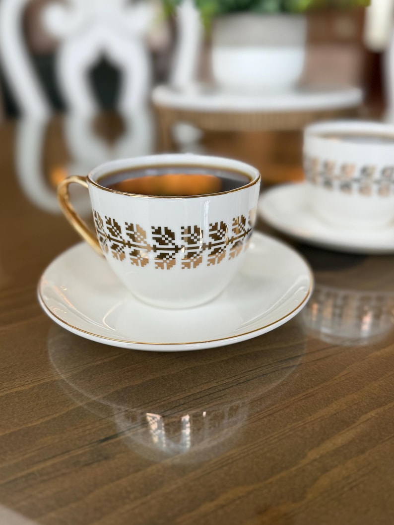 May include: A white teacup and saucer set with gold trim and a geometric gold pattern. The cup is filled with dark liquid, likely coffee or tea. The set is on a reflective wooden surface, with a second cup and saucer visible in the background.