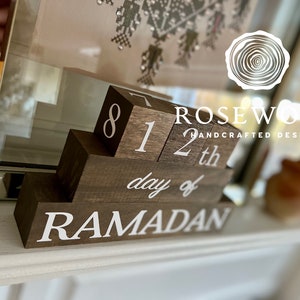 May include: A wooden Ramadan calendar with the text "8 12th day of RAMADAN" in white. The calendar is made of dark brown blocks and is designed to be displayed on a shelf or mantel. The brand name "Rosewood Handcrafted Designs" is also visible.