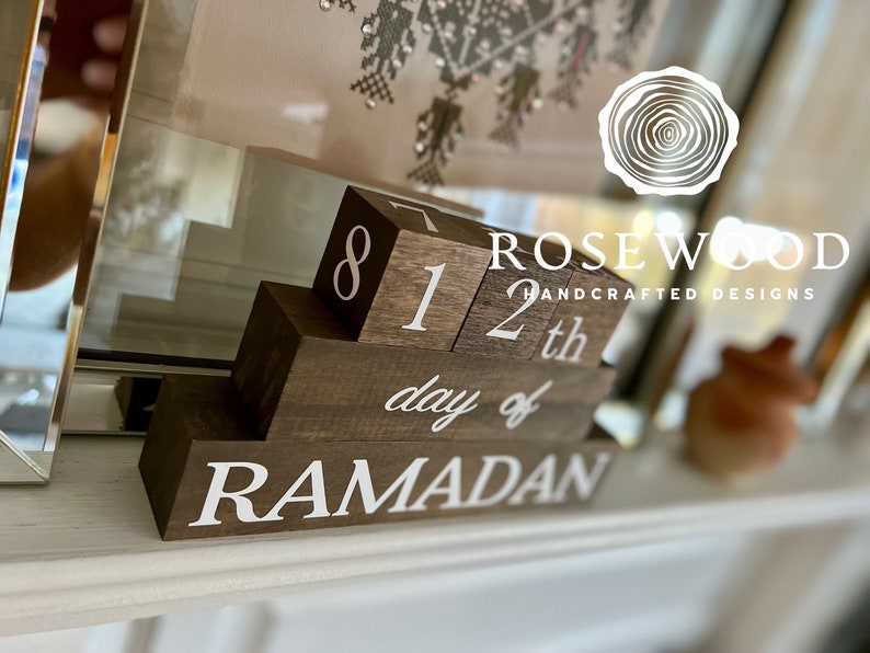 May include: A wooden Ramadan calendar with the text "8 12th day of RAMADAN" in white. The calendar is made of dark brown blocks and is designed to be displayed on a shelf or mantel. The brand name "Rosewood Handcrafted Designs" is also visible.