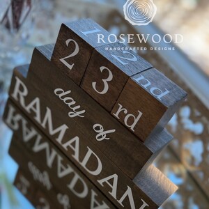 May include: A wooden block calendar with the text "day of Ramadan" and numbers 1, 2, 3, and 22, 23, and "nd" in white. The blocks are arranged in a staggered fashion. The brand name "Rosewood Handcrafted Designs" is also visible.