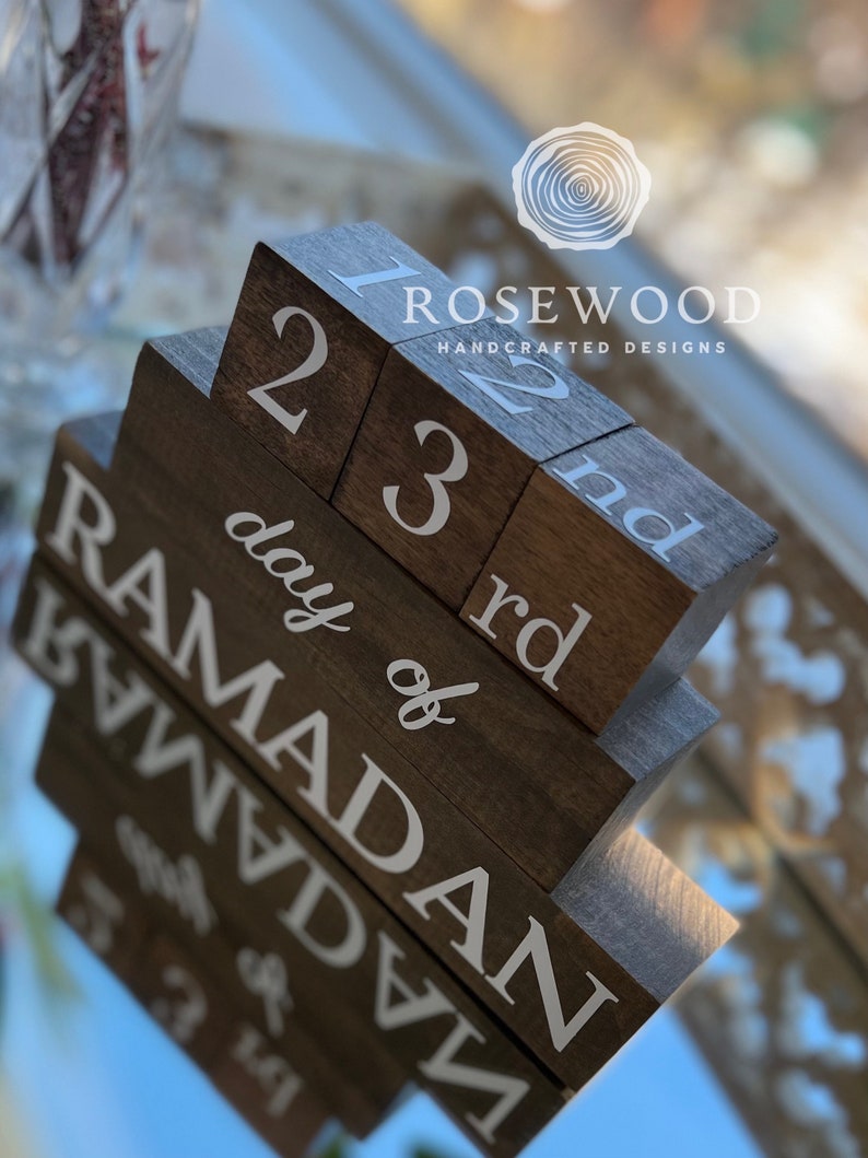 May include: A wooden block calendar with the text "day of Ramadan" and numbers 1, 2, 3, and 22, 23, and "nd" in white. The blocks are arranged in a staggered fashion. The brand name "Rosewood Handcrafted Designs" is also visible.