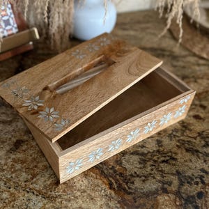 May include: A wooden tissue box with a hinged lid, featuring inlaid floral designs. The box is made of light-colored wood and has a rectangular shape. The lid is partially open, revealing the interior. The floral accents are silver-colored.