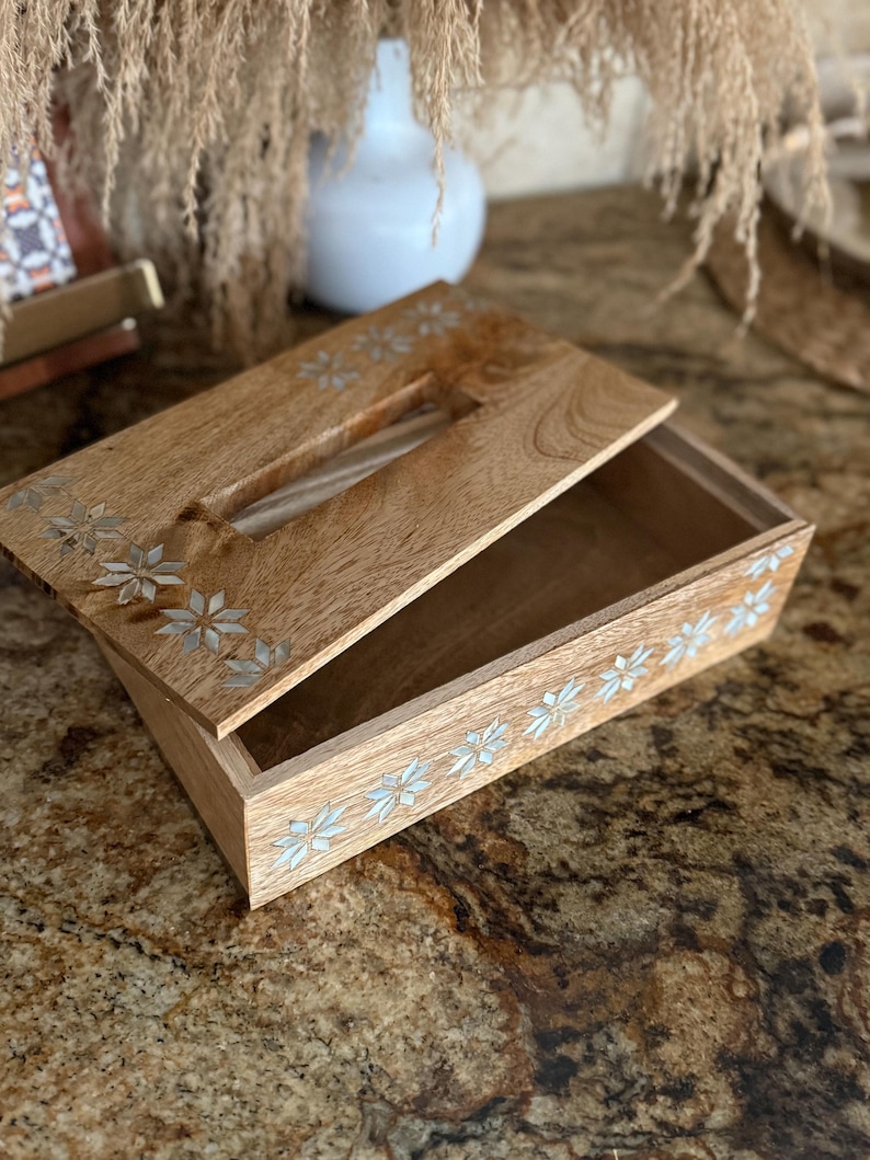 May include: A wooden tissue box with a hinged lid, featuring inlaid floral designs. The box is made of light-colored wood and has a rectangular shape. The lid is partially open, revealing the interior. The floral accents are silver-colored.
