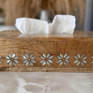 May include: Wooden tissue box with a decorative inlaid star pattern along the front edge. The box is made of light brown wood and contains white tissues. The star pattern is made of a mother-of-pearl material.