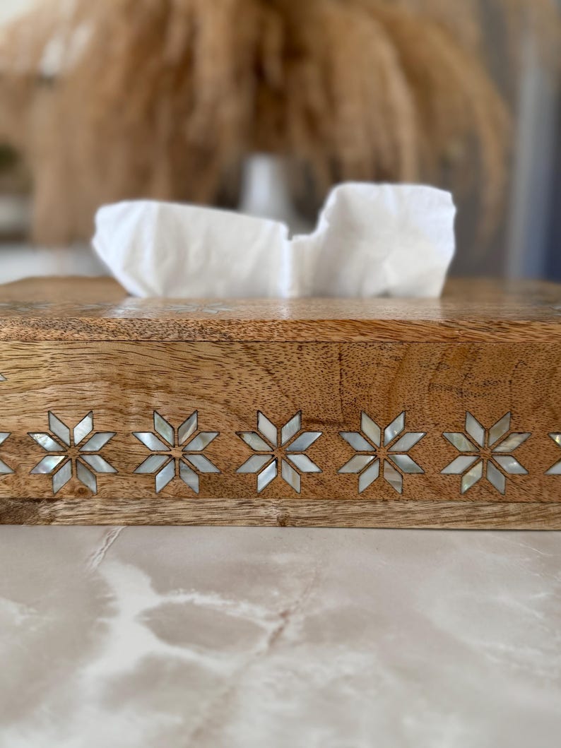 May include: Wooden tissue box with a decorative inlaid star pattern along the front edge. The box is made of light brown wood and contains white tissues. The star pattern is made of a mother-of-pearl material.