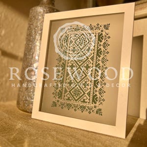 K&ouml;nnte beinhalten: Eine gerahmte Kreuzstich-Stickerei mit einem gr&uuml;nen geometrischen Muster auf beigefarbenem Hintergrund. Der Rahmen ist wei&szlig; und der Text "ROSEWOOD HANDCRAFTED GIFTS DECOR" ist unten sichtbar.