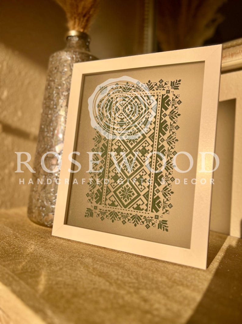 K&ouml;nnte beinhalten: Eine gerahmte Kreuzstich-Stickerei mit einem gr&uuml;nen geometrischen Muster auf beigefarbenem Hintergrund. Der Rahmen ist wei&szlig; und der Text "ROSEWOOD HANDCRAFTED GIFTS DECOR" ist unten sichtbar.