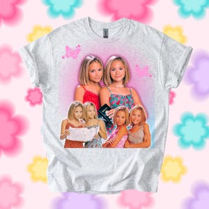 Pu&ograve; includere: T-shirt grigio chiaro con una grafica di due donne. La grafica ha uno sfondo rosa con farfalle e pi&ugrave; immagini delle donne. La t-shirt &egrave; uno stile girocollo.