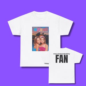 May include: White t-shirt featuring a movie poster graphic of two young women with the text "OUR LIPS ARE SEALED" and the text "mary-kate and ashley" on the front. The back of the shirt has the word "FAN" in bold black letters.