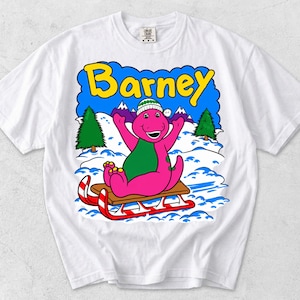 Op de afbeelding: Een wit t-shirt met korte mouwen en een grafische print van de paarse dinosaurus Barney met een groene sjaal en een wit-groene muts, zittend op een bruine slee in de sneeuw met groene dennenbomen en de gele tekst Barney erboven.
