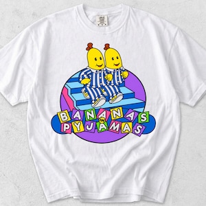 May include: A white short-sleeve t-shirt featuring a graphic of two yellow banana characters wearing blue and white striped pajamas. The characters are walking down blue stairs inside a purple circle. The text reads BANANAS IN PYJAMAS.