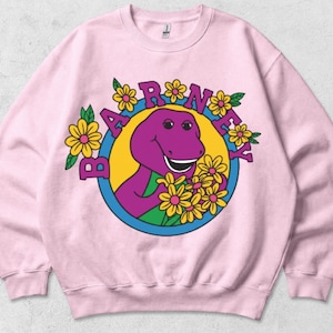 Puede incluir: Una sudadera de cuello redondo rosa claro con un estampado gráfico de un personaje de dinosaurio morado sosteniendo flores amarillas. El diseño está encerrado en un círculo azul con el texto BARNEY arqueado encima, rodeado de flores y hojas.