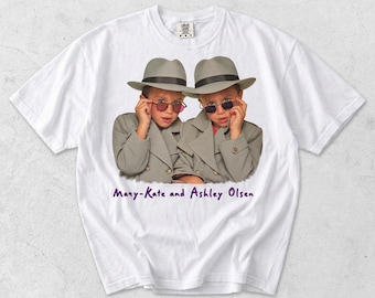 May include: A white short-sleeved t-shirt featuring a graphic of two children wearing grey trench coats, grey fedoras, and sunglasses. Below the graphic, the text reads "Many-Kate and Ashley Olsen" in purple script. The shirt has a crew neckline.
