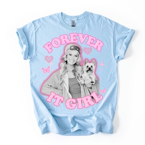 May include: Light blue t-shirt with the text "FOREVER IT GIRL" in pink bubble letters. The shirt features a black and white image of a woman holding a small dog, with pink hearts and butterflies.