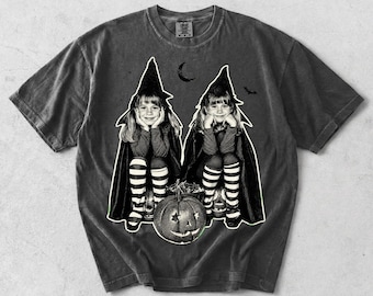 May include: A charcoal gray short-sleeve t-shirt featuring a black and white graphic of two children in witch costumes with pointed hats and striped socks. They are sitting behind a carved jack-o'-lantern with star-shaped eyes under a crescent moon.
