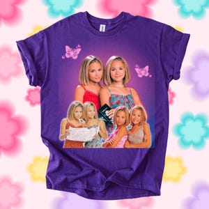 Pu&ograve; includere: T-shirt viola con una grafica di due donne bionde con top colorati. La grafica include farfalle e immagini aggiuntive delle donne. Girocollo.