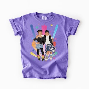 May include: A lilac t-shirt with a graphic of two children on roller skates, set against a backdrop of colourful starbursts and stars. The image has a vintage, retro aesthetic.