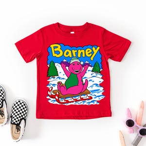 Op de afbeelding: Een felrood T-shirt met korte mouwen en een afbeelding van de paarse dinosaurus Barney met een groen-witte wintermuts op een houten slee in de sneeuw. De tekst Barney staat in het geel boven het personage met dennenbomen op de achtergrond.