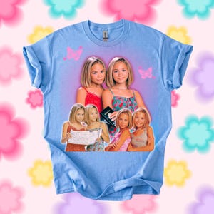 Pu&ograve; includere: T-shirt azzurra con una grafica di due giovani donne, con immagini aggiuntive delle stesse donne. Sono presenti anche grafiche di farfalle rosa. La maglietta ha maniche arrotolate e girocollo.