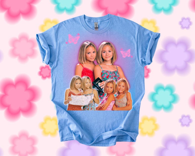 Pu&ograve; includere: T-shirt azzurra con una grafica di due giovani donne, con immagini aggiuntive delle stesse donne. Sono presenti anche grafiche di farfalle rosa. La maglietta ha maniche arrotolate e girocollo.