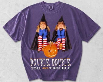 May include: A purple short-sleeve t-shirt featuring a graphic of two children dressed as witches with black capes and hats, red and white striped socks, and a carved orange pumpkin. The text reads: DOUBLE, DOUBLE, TOIL AND TROUBLE.