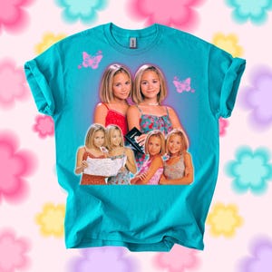 Pu&ograve; includere: T-shirt color turchese con una grafica di due donne, con immagini pi&ugrave; piccole delle stesse donne. Sullo sfondo ci sono farfalle rosa. La maglietta &egrave; del marchio Gildan.