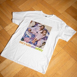Puede incluir: Camiseta blanca con una imagen retro de dos personas en pijama en una cama. El texto "mary-kateandashley" está impreso en naranja y verde. La camiseta tiene una estética vintage.