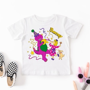 Puede incluir: Una camiseta blanca de manga corta con un gráfico colorido del dinosaurio morado Barney con un gorro de fiesta, un niño con un vestido rosa de lunares soplando un matasuegras y un perro pequeño, con el texto Barney y estrellas amarillas.