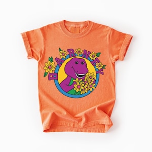 Puede incluir: Una camiseta naranja de manga corta con un gráfico del dinosaurio morado Barney rodeado de flores amarillas dentro de un círculo azul. La palabra BARNEY está impresa en letras mayúsculas moradas arqueadas sobre el diseño circular.
