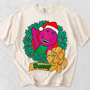 Puede incluir: Una camiseta de manga corta color crema con un gráfico del dinosaurio morado Barney usando un gorro de Papá Noel rojo y blanco. Barney está centrado dentro de una corona de acebo verde con bayas rojas, acentuada por un gran lazo dorado y el texto Barney.