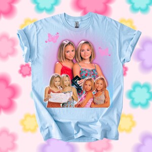 Pu&ograve; includere: T-shirt azzurra con una grafica di due giovani donne in abiti colorati. Farfalle rosa sono stampate sulla maglietta. La maglietta ha maniche arrotolate e girocollo.