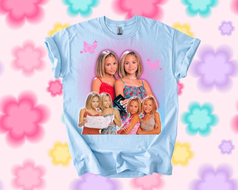 Pu&ograve; includere: T-shirt azzurra con una grafica di due giovani donne in abiti colorati. Farfalle rosa sono stampate sulla maglietta. La maglietta ha maniche arrotolate e girocollo.