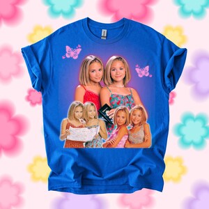 Pu&ograve; includere: T-shirt blu reale con una grafica di due giovani donne, con farfalle. La grafica include pi&ugrave; immagini delle donne in vari outfit. La maglietta &egrave; del marchio Gildan.
