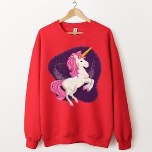 Op de afbeelding: Rode sweatshirt met een wit eenhoornontwerp, roze manen en staart, en gouden hoorn. De eenhoorn staat tegen een paarse achtergrond met een gestikte rand. De sweatshirt heeft lange mouwen en een ronde hals.