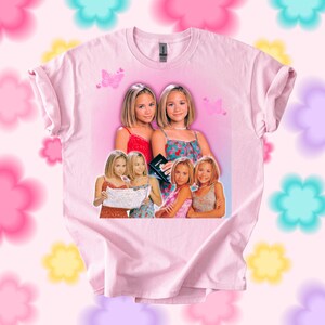 Pu&ograve; includere: T-shirt rosa chiaro con una grafica di due giovani donne in vari outfit. La grafica include farfalle e uno sfondo rosa. La maglietta &egrave; a girocollo.