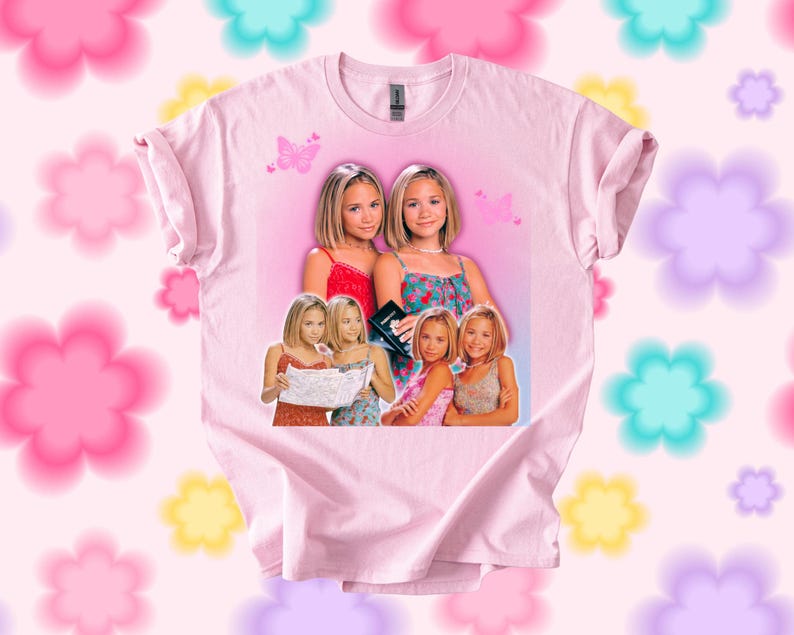 Pu&ograve; includere: T-shirt rosa chiaro con una grafica di due giovani donne in vari outfit. La grafica include farfalle e uno sfondo rosa. La maglietta &egrave; a girocollo.