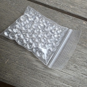 May include: A clear plastic bag filled with numerous transparent, spherical glass marbles. The bag is sealed with a zipper closure. The marbles are densely packed inside the bag, creating a visually interesting texture. The bag is resting on a wooden surface.