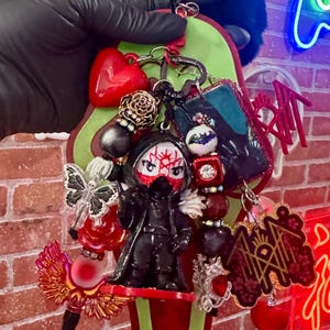 May include: A decorative keychain featuring a central hooded figure with a white and red mask, surrounded by various charms including a red heart, a butterfly with a skeleton print, a rectangular image, and a dark red logo charm with gold text.