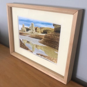 May include: Framed landscape photograph featuring rock formations reflected in water under a blue sky. The photograph is set within a light wood frame with a cream-colored mat.