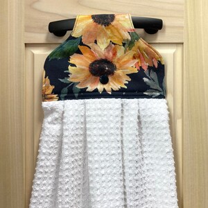 May include: A hanging kitchen towel with a sunflower design. The top portion features a black background with yellow and orange sunflowers and green leaves. The bottom is a white, textured towel, and the towel is attached to a black bar.