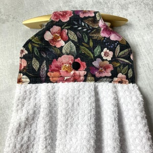May include: A white kitchen towel with a floral print hanging on a gold hook. The floral print features pink, purple, and white flowers on a black background.