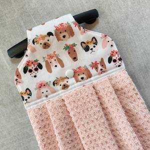 May include: A pink and white kitchen towel with a hanging loop and a button. The towel features a white fabric with a pattern of dogs wearing floral crowns.