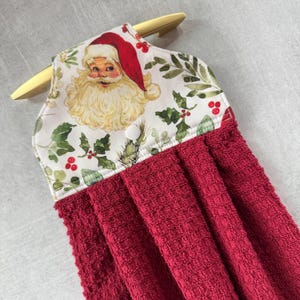 May include: A red and white Christmas themed kitchen towel with a Santa Claus print. The towel has a loop for hanging and a button.