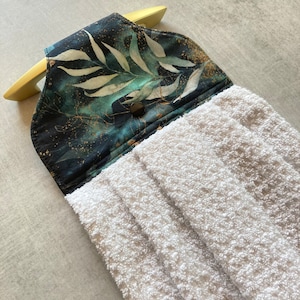 Peut inclure: Une serviette à main blanche et texturée avec un dessus décoratif orné d'un motif botanique dans les tons de vert d'eau, or et noir. La serviette est conçue pour être suspendue à une poignée dorée.