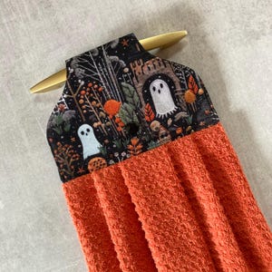 May include: Orange Halloween-themed kitchen towel with a black top featuring a spooky forest scene with ghosts and autumn foliage. The towel is designed to hang from a gold-colored handle, adding a festive touch to any kitchen.