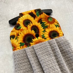 May include: A hanging kitchen towel with a sunflower pattern on the top portion. The towel has a gray, textured bottom and a black snap closure. The sunflower design features yellow petals and dark brown centers.