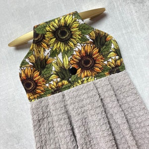 May include: A gray kitchen towel with a sunflower print hanging loop. The loop is made of white fabric with a sunflower print. The towel is made of a soft, absorbent material.