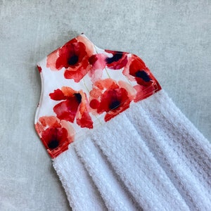 May include: White kitchen towel with a red poppy floral print on the top. The towel is made of terry cloth and has a hanging loop.